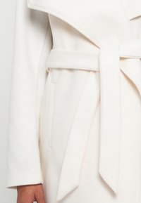 White wool coat with a wide collar, tied belt, and side pockets. Smooth texture with a clean, minimal design.