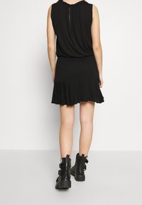 Kobieta w czarnej sukience bez rękawów i czarnych butach za kostkę z sznurowaniem, stojąca tyłem na tle gładkiego białego tła.