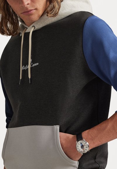 Sweatshirt cinza escuro com um bolso cinza claro, mangas azuis e capuz cinza claro. Apresenta um cordão e logótipo bordado. Relógio de pulso preto visível.