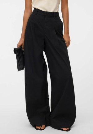 Femme portant un pantalon noir taille haute à jambes larges, top beige, tenant une pochette noire pliée, debout avec les pieds dans des sandales noires.