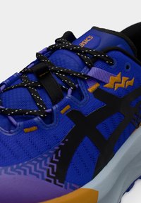 Μπλε αθλητικό παπούτσι με επάνω μέρος από δίχτυ, μαύρες λεπτομέρειες, υφή και φωτεινές πορτοκαλί και μοβ αποχρώσεις. Μαύρα κορδόνια συμπληρώνουν το σχέδιο.