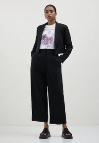 Frau steht mit den Händen in den Taschen, trägt ein schwarzes, kurzes Blazer, weite Hose, ein weißes T-Shirt mit floralem Grafikdesign und schwarze Plateausandalen.