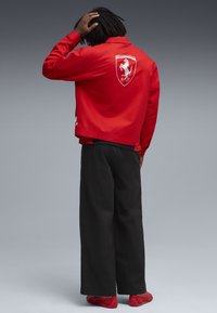 Jachetă roșie cu un logo alb Ferrari pe spate, asortată cu pantaloni negri evazați și pantofi roșii. Fundalul este un gradient gri.