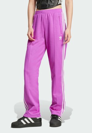 Person iført lyse lilla Adidas træningsbukser med hvide striber og sorte Adidas sneakers med hvide striber, stående mod en lys baggrund.