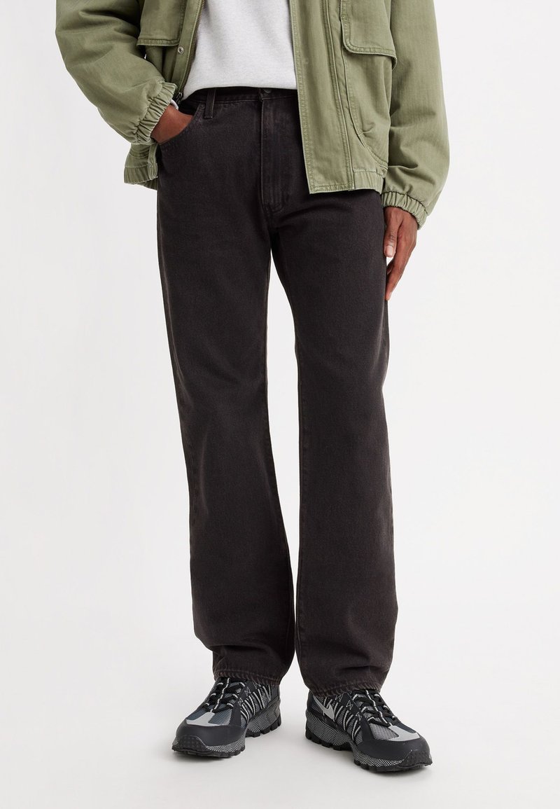 Donkergrijze rechte jeans van katoen, gecombineerd met een lichtgroene bomberjack en zwarte gestructureerde sneakers. Focus op casual outfit.