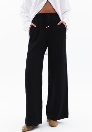 Person trägt lockere schwarze Hose mit Kordelzug in der Taille und hat die Hände in den Taschen, kombiniert mit hellfarbenen gewebten Schuhen und einem weißen Oberteil.