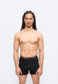 Μυώδης άνδρας με μακριά dreadlocks στέκεται μπροστά, φοράει μαύρα μποξεράκια Calvin Klein σε απλό λευκό φόντο.