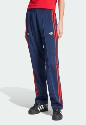 Osoba nosząca granatowe spodnie dresowe Adidas z czerwonymi paskami i logo, w połączeniu z czarnymi butami Adidas z białymi paskami.