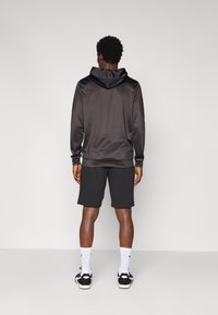 Czarna sportowa bluza z kapturem o teksturowanym wykończeniu, zestawiona z dopasowanymi czarnymi szortami. Model stoi na tle jednolitego jasnego tła.