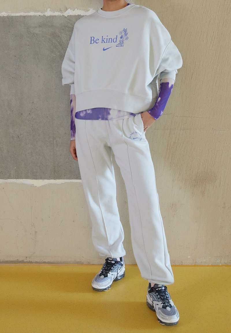 Ljust grå, oversize sweatshirt med texten "Var snäll" och en blommig grafik, i kombination med matchande sweatpants. Har långa ärmar i lila tie-dye.