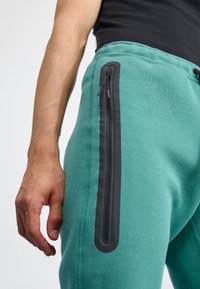 Pantaloni tuta turchesi realizzati in morbido tessuto, con una tasca con zip nera lungo la coscia esterna e vita regolabile con cordino.