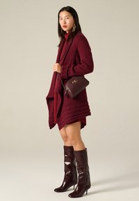 Cardigan lavorato a maglia bordeaux con una vestibilità morbida, indossato sopra un vestito coordinato, abbinato a stivali alti fino al ginocchio bordeaux lucidi e a una piccola borsa.