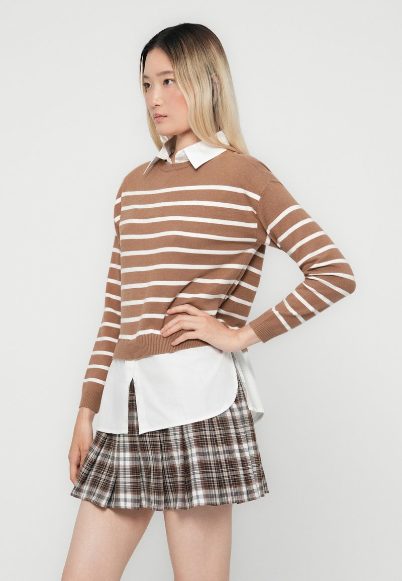Maglione a righe marroni e bianche indossato sopra una camicia bianca con bottoni, abbinato a una mini gonna a quadri in tonalità marroni, bianche e grigie.