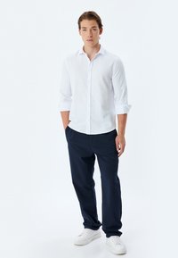 Camisa blanca de botones con mangas remangadas, combinada con pantalones azul marino y zapatillas blancas. Tela suave y diseño minimalista.