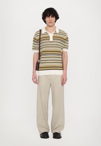 Polo a righe a maniche corte nei colori marrone, crema e verde, con colletto bianco; abbinato a pantaloni a gamba larga beige chiaro e scarpe nere.
