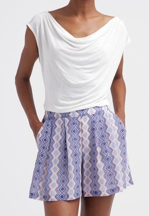 Frau trägt ein weißes, drapiertes ärmelloses Oberteil kombiniert mit High-Waist-Shorts mit einem blauen und pfirsichfarbenen geometrischen Muster.