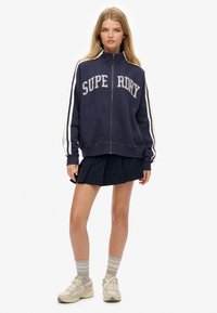 Sudadera con cremallera en color navy con el texto "SUPERDRY", rayas blancas en las mangas, combinada con una falda navy plisada y zapatillas beige. Tejido texturizado.
