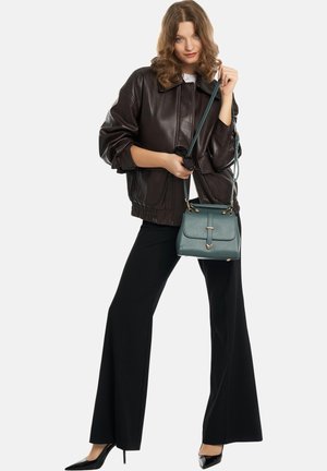 Vrouw draagt een donkerbruine leren jas, zwarte uitlopende broek, zwarte hakken, en houdt een groene handtas bij het hengsel vast.