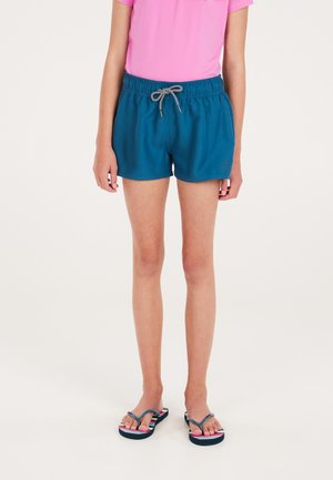 Jong persoon draagt turquoise elastische shorts met witte trekkoord en roze top, staand op donkerblauwe slippers tegen een effen achtergrond.