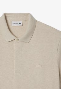 Beige pikétröja med en strukturerad tyg, en mjuk krage och en knappslå med tre knappar. Har Lacoste-logotyper broderade på bröstet.