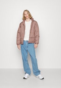 Giacca marrone imbottita con colletto alto, indossata sopra una camicia bianca. Jeans azzurri larghi e scarpe da ginnastica bianche completano l'outfit.