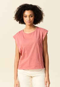 Lyhyt hiha, löysä t-paita pehmeästä, pölyisen vaaleanpunaisesta puuvillasta, jossa on pyöreä kaula-aukko ja hillityt hihansuut, yhdistettynä vaaleisiin pantsiin.