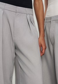 Deux personnes portant un pantalon plissé gris clair avec des passants de ceinture, une main reposant sur le côté, toutes deux avec un haut rentré.