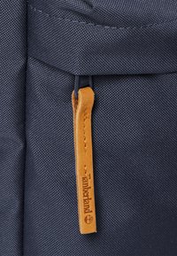 Námořnický látkový batoh s texturovaným povrchem, se zipem na koženém táhle a jemným brandingem. Všimněte si tmavého zipového hardwaru.