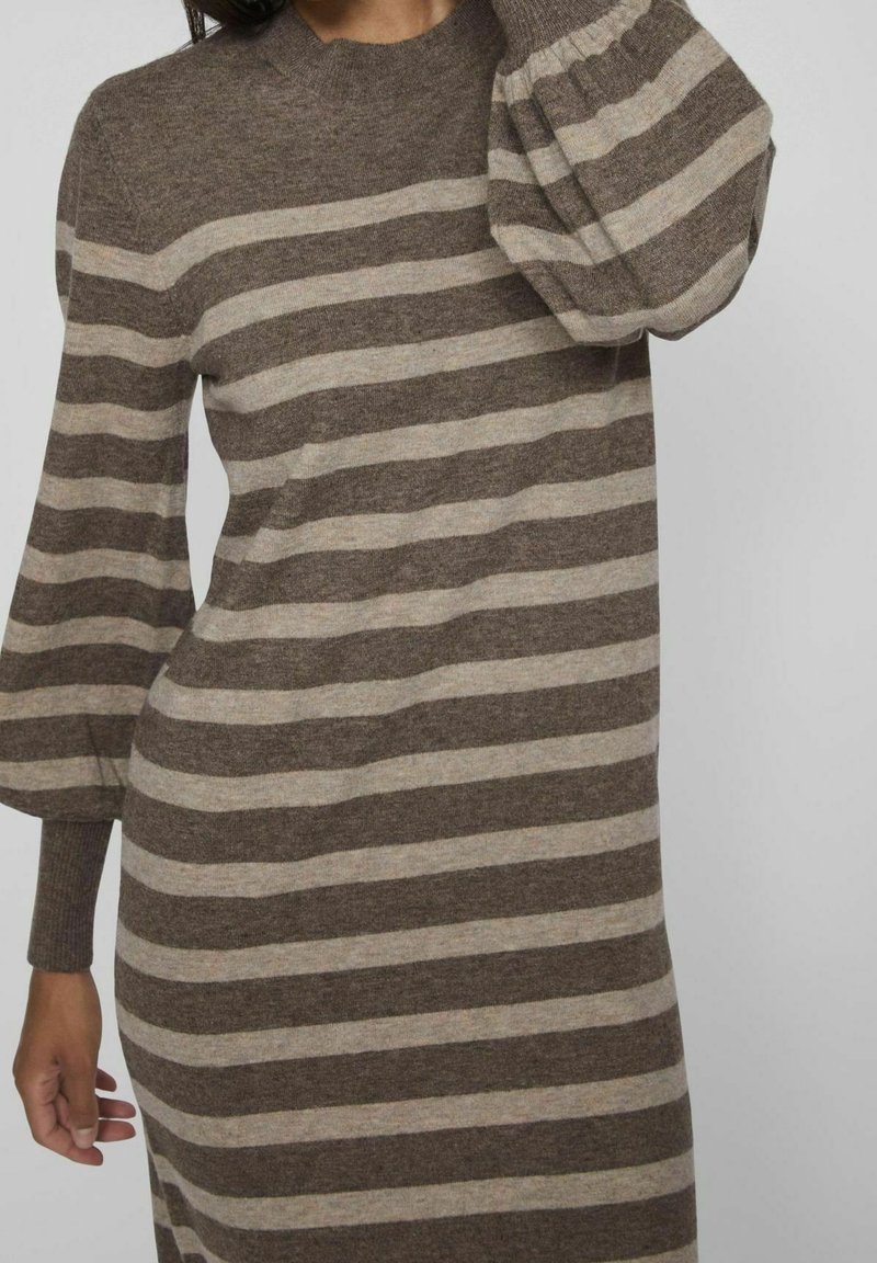 Femme portant une robe tricotée à rayures horizontales marron et beige avec des manches longues bouffantes et des poignets ajustés.