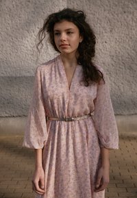 Robe florale rose avec de longues manches bouffantes, décolleté en V, taille cintrée avec une ceinture en argent, tissu texturé et motifs subtils violets.