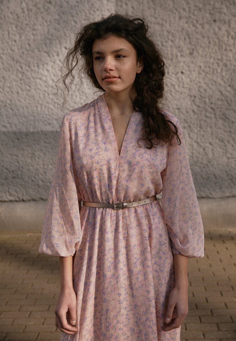 Robe florale rose avec de longues manches bouffantes, décolleté en V, taille cintrée avec une ceinture en argent, tissu texturé et motifs subtils violets.