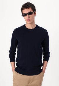 Pull marin bleu à col rond et manches longues, fabriqué à partir d'un matériau doux et texturé. Porté avec des lunettes de soleil foncées, associé à un pantalon beige.