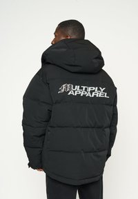 Giacca a vento nera con cappuccio, caratterizzata da un design trapuntato e la scritta "MULTIPLY APPAREL" in bianco sul retro.