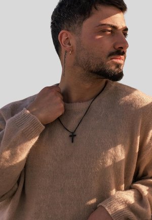 Beige knitted sweater with a round neck. Black cross pendant on a thin necklace. Model has short hair and a visible tattoo on the neck.