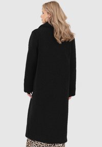 Cappotto teddy nero con maniche lunghe e colletto. Tessuto morbido e testurizzato, lunghezza al ginocchio, indossato sopra una gonna a fantasia.