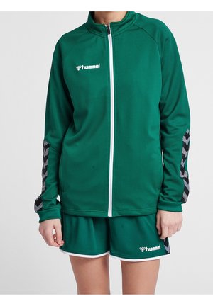 Persoon draagt groene hummel sportkleding met een wit jack met rits en bijpassende korte broek, voorzien van chevronpatronen op de mouwen.