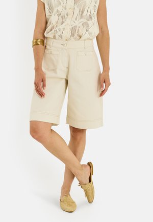 Personne portant un short beige longueur genou, un chemisier sans manches à motifs, un bracelet doré et des chaussures beiges tressées sans lacets, debout les jambes croisées.