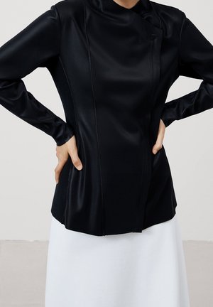 Kobieta w dopasowanej czarnej bluzce z długim rękawem, z ukośnymi przeszyciami, oraz białej spódnicy, z rękami opartymi na biodrach na jednolitym tle.