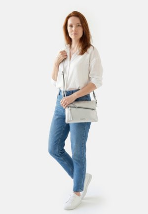 Woman with red hair wearing white shirt, blue jeans, and white sneakers, holding a silver crossbody purse over her shoulder.