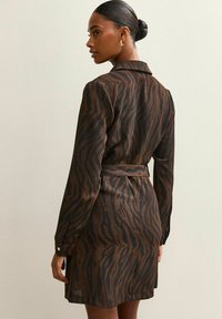 Mujer con el cabello oscuro recogido en un moño, vistiendo un vestido con cinturón de estampado de cebra en tonos marrón y negro, de perfil contra una pared lisa de color claro.