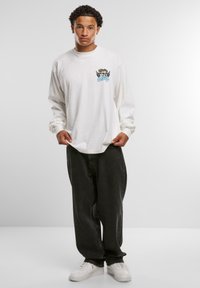 Hvid langærmet T-shirt med et blåt grafisk design med en fisk og vinger på brystet, parret med løse sorte jeans og hvide sneakers.
