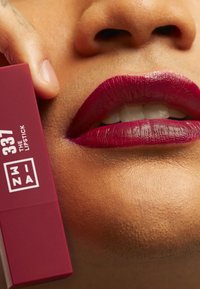 Burgundy läppstift i ett rektangulärt, bordeauxfärgat etui som visar "337 THE LIPSTICK" och en vit logotyp, hålls nära fylliga läppar med en glansig finish.