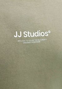 Alyvuogių žalios spalvos medvilninė medžiaga su lygiu paviršiumi. Baltas atspaustas logotipas: „JJ Studios“, po juo mažesniu šriftu nurodyta dizaino kilmė.