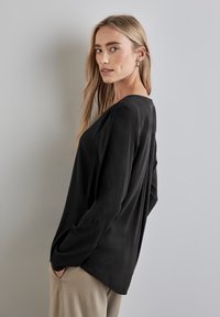 Blouse noire à manches longues avec une coupe décontractée, présentant un col rond, des plis subtils aux épaules et des poignets boutonnés. Texture lisse.