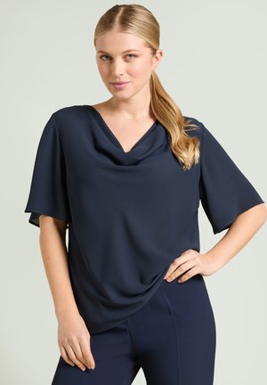 Kvinde med blond hestehale iført løs, marineblå bluse og matchende bukser, stående med den ene hånd på hoften mod en lys baggrund.