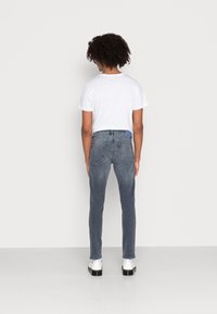 Vaqueros pitillo gris oscuro con cinco bolsillos, textura desgastada y etiqueta azul en la parte trasera. Combinados con una camiseta blanca lisa y zapatos negros.