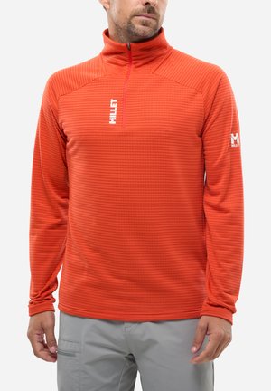 Man wearing a bright orange long-sleeve zip-up pullover with "Millet" logo on chest and left sleeve, paired with light gray pants.