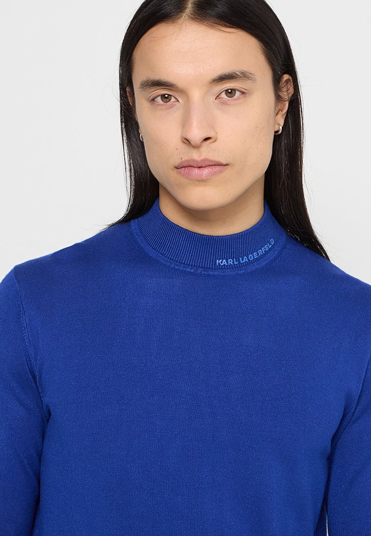 Blauer gestrickter Pullover mit Mock-Neck, der einen gerippten Kragen und dezente Markenkennzeichnung am Ausschnitt aufweist. Glatte Textur mit figurbetontem Schnitt.