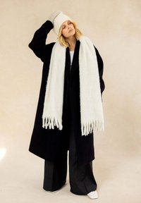 Manteau long noir, écharpe blanche et duveteuse avec franges, bonnet en tricot crème, haut blanc, pantalon à rayures larges, et chaussures blanches. Fond neutre.
