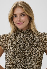Animal-print blouse in beige and brown featuring a high ruffled neckline, decorative buttons, and soft, sheer texture with frill details.